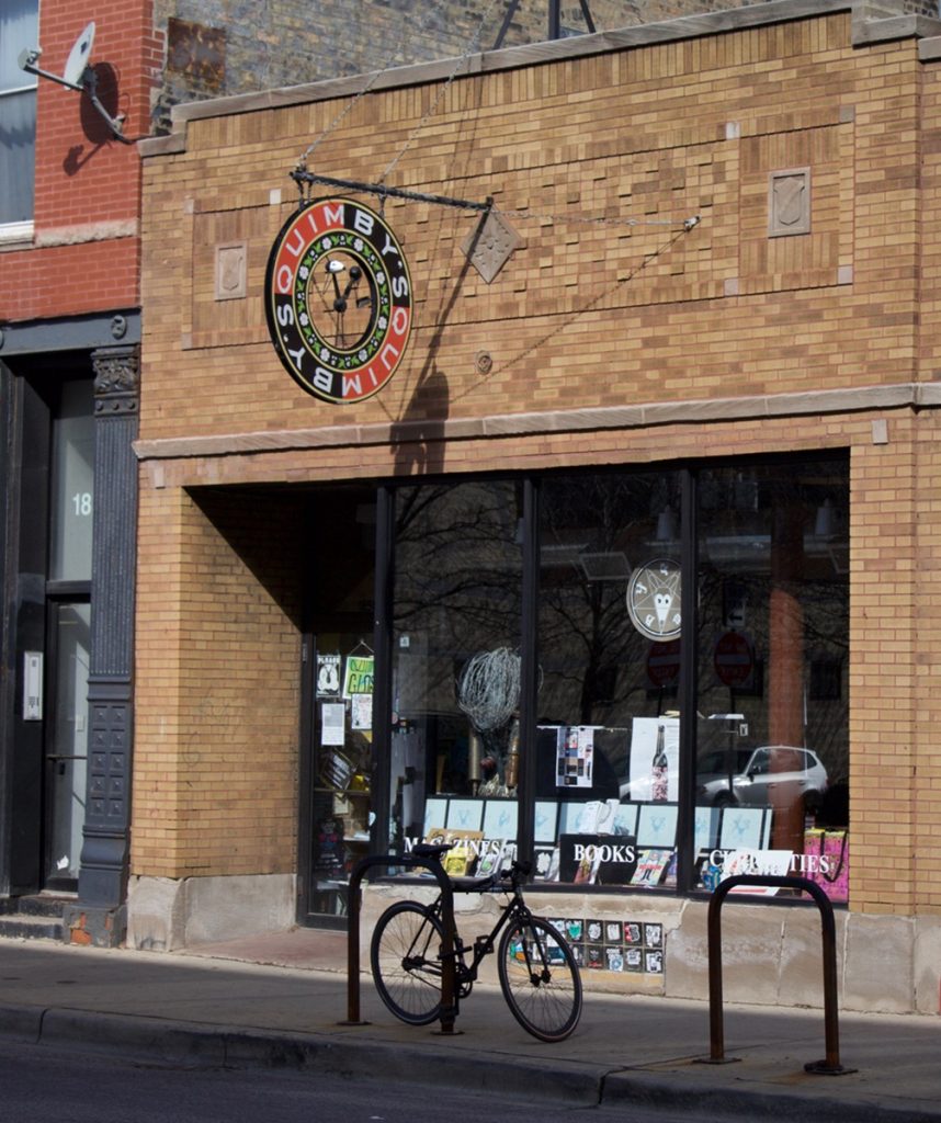 A zoomed-out photo of the outside storefront for Quimby's Bookstore in Chicago, with the red and black circuclar signage hanging above the dark windowed space. An apartment entrance is just out of view on the leftside, with the stark reflection of the opposite street in the store windows above the displays of zines and comics.