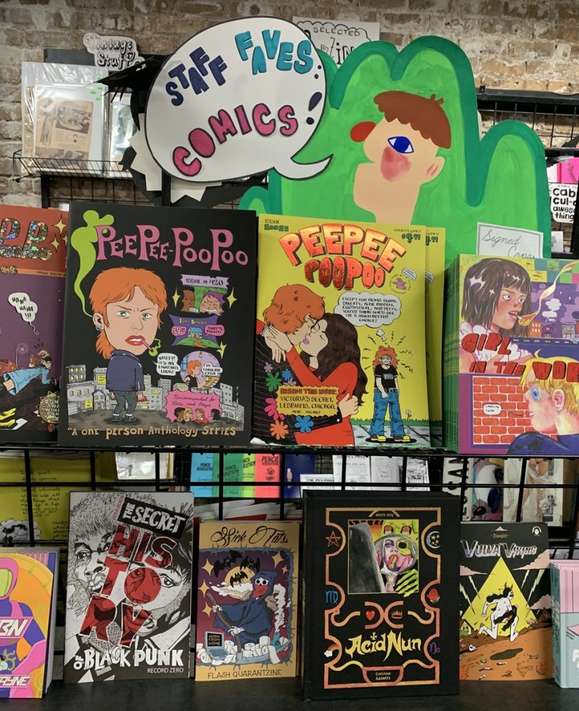 A staff favorites shelf at Quimby's Bookstore, with a green paper cutout on top. On the black wire rack, Silver Sprocket comics are displayed, including (from left to right) PeePee PooPoo issues #69, 420, 80085, Girl in the World, LSBN, The Secret History of Black Punk, Sick Tats, Acid Nun, Fungirl: Vulva Viking and Fart School