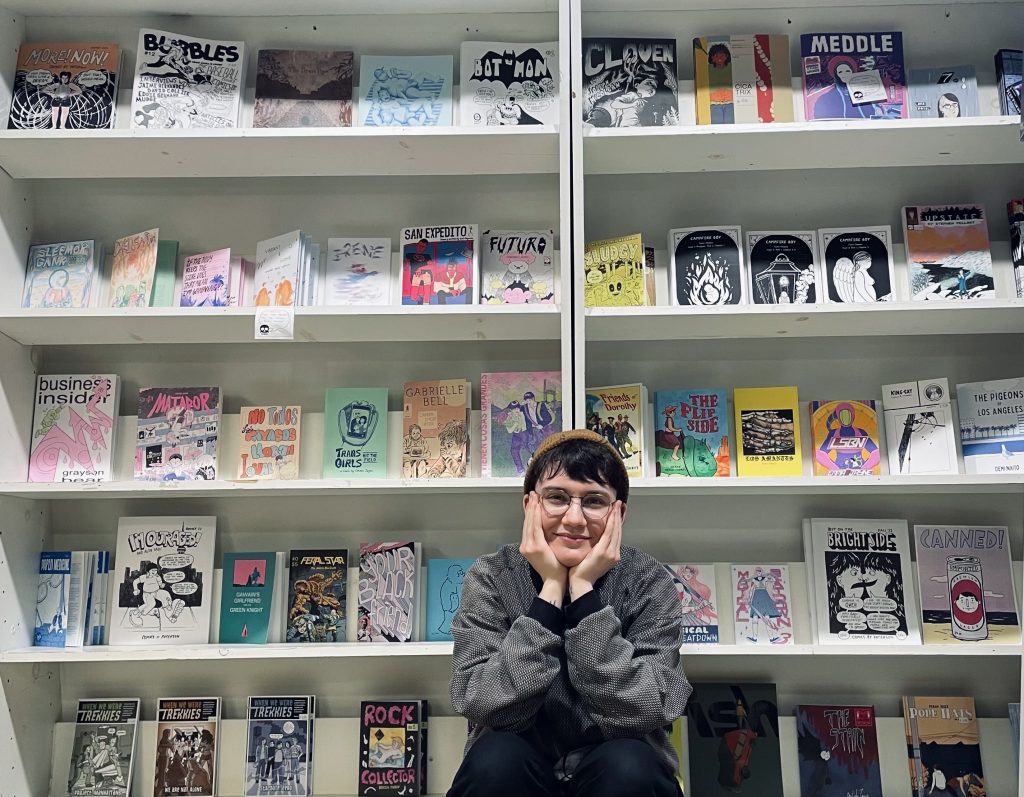A smiling Vault of Midnight bookseller with hands clasped under their face, sitting in front of two large shelves of small press comics.