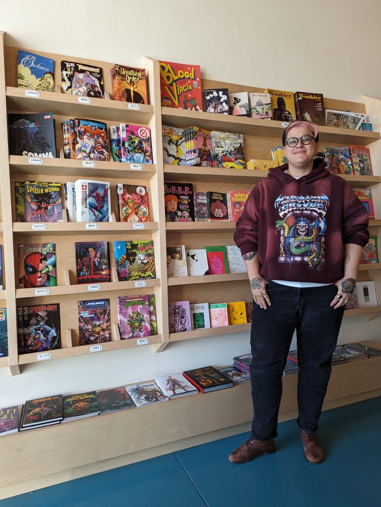 A photo of Secret Headquarters manager Jules in front of shelves of single issue comics streching out behind them. Jules has pink hair, round glasses, a red Cheatin Snakes hoodie on with some brown boots and black jeans. 