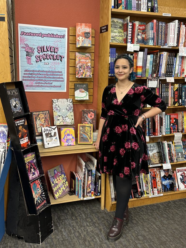 Mo smiling, standing to the right of their Silver Sprocket comic display and in front of a larger comic and graphic novel section.
