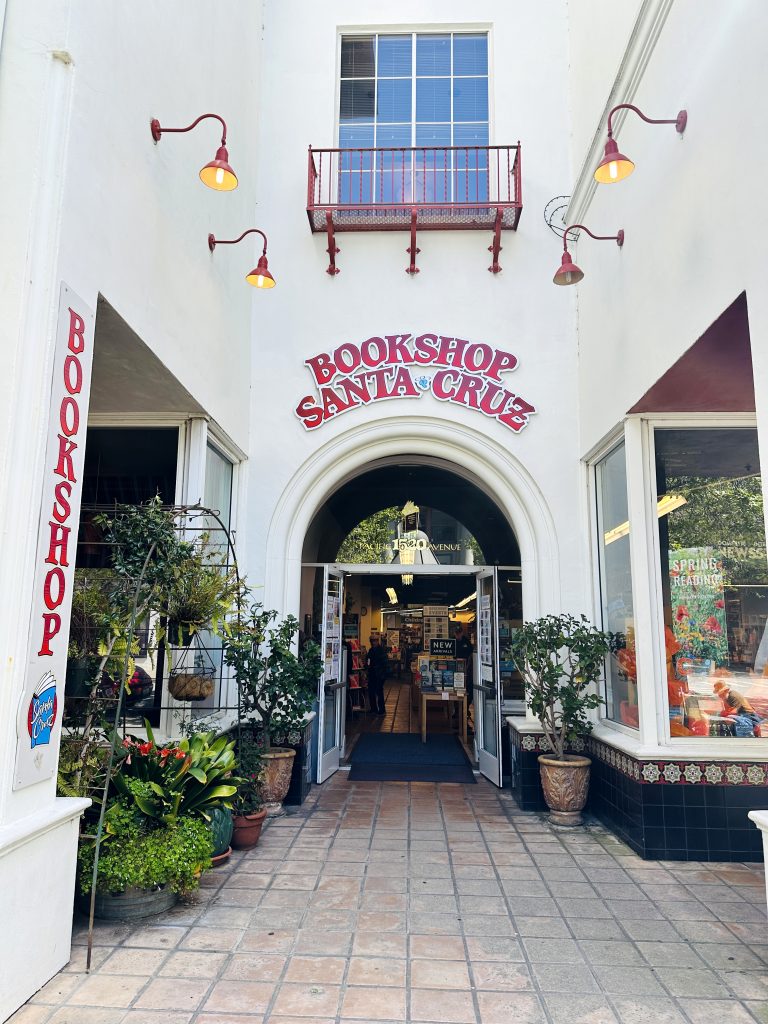 Outside storefront for Bookshop Santa Cruz. Assorted plants cover the left side with the open doors centered to show a glimpse at the inside of the shop.