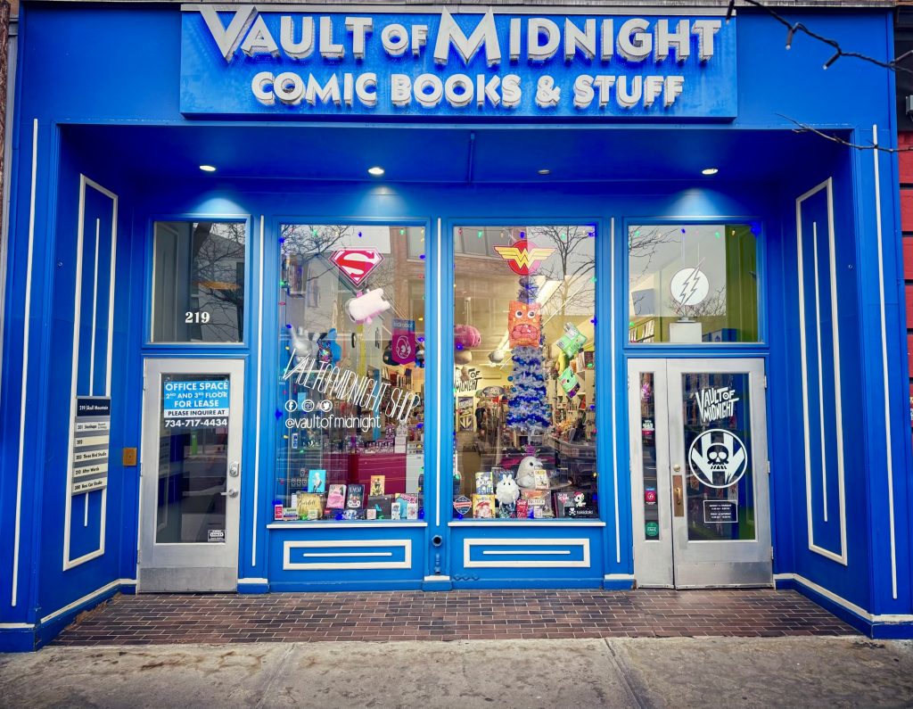 The outside storefront of Vault of Midnight with it's deep blue awning and big white block letters over the entrance. Blue and white accents square accents surround tall store windows with comics and superhero logos covering them. On the right side, you can see the door with the text Vault of Midnight and the skull logo printed on the glass.