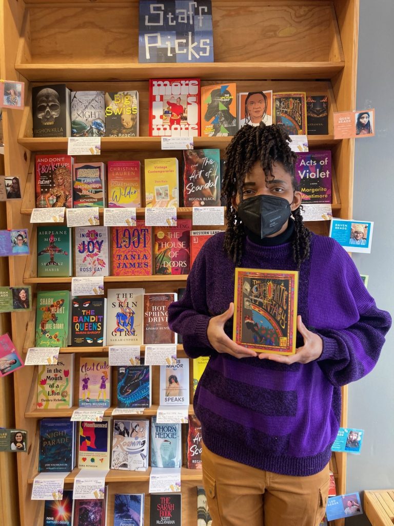 Bookseller Quen standing in front of a Staff Picks section, holding The Chromatic Fantasy in front of her.