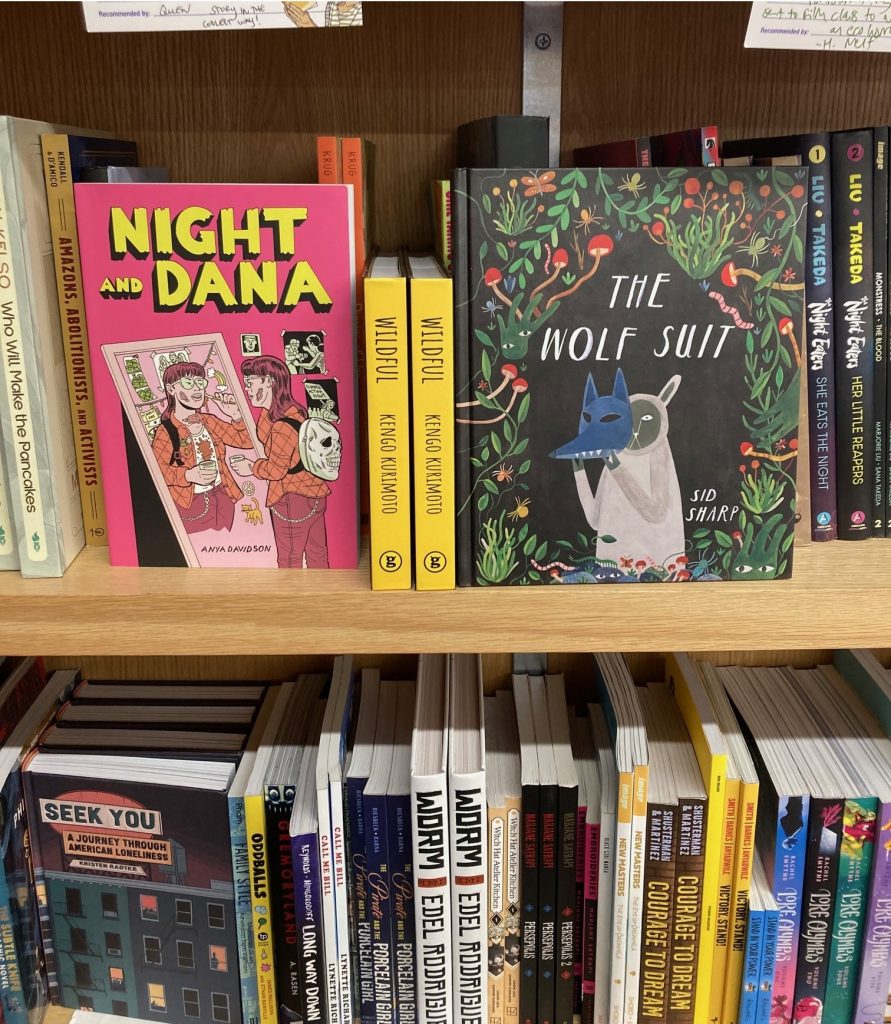 The books "Night and Dana" by Anya Davidson and "The Wolf Suit" by Sid Sharp, facing out on a wooden shelf filled with other comics.