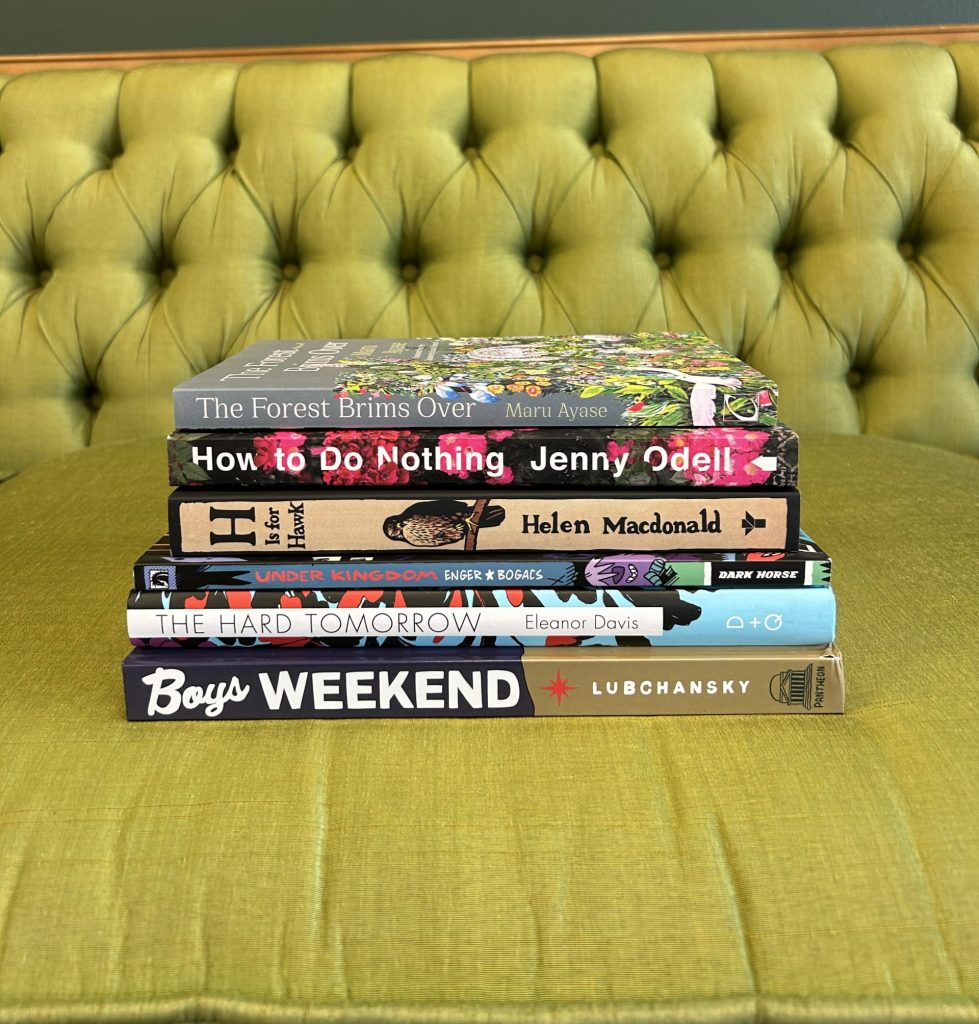 6 book stack centered on an plush couch, including Boys Weekend, The Hard Tomorrow, Under Kingdom, H is for Hawk, How to Do Nothing, and The Forest Brims Over.